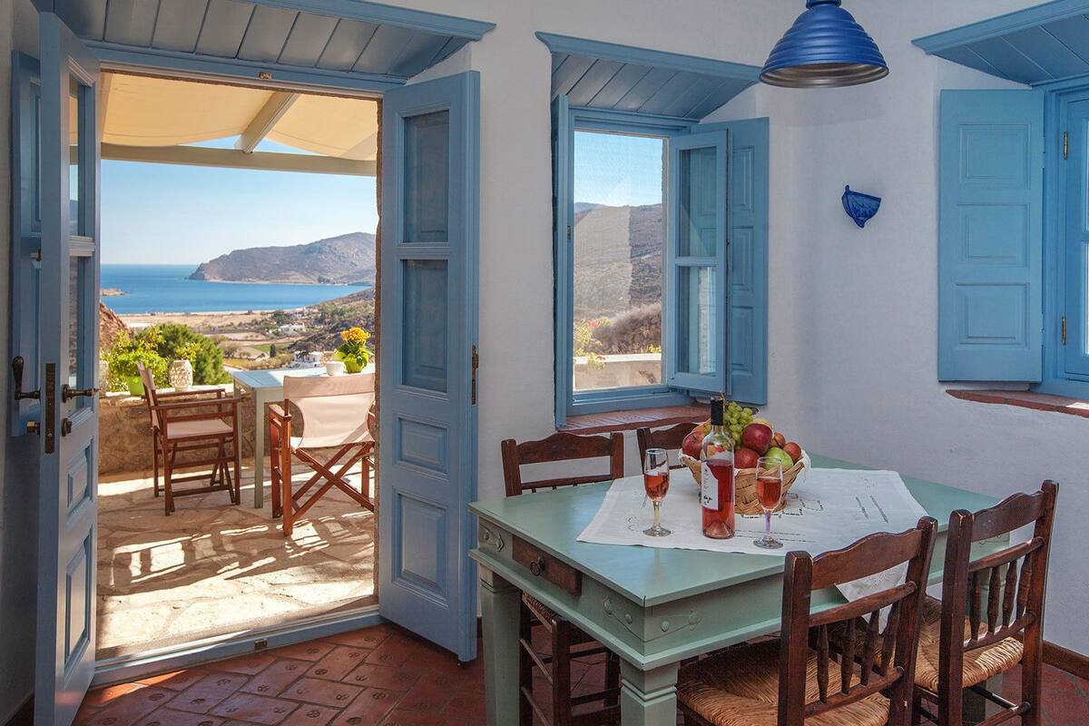 An open door leading to a terrace with a table set for two, a bottle of wine, and a basket of fruit.