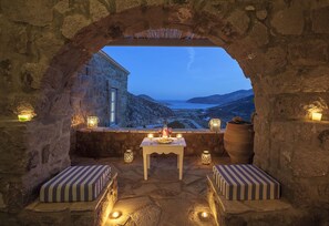 A stone-walled outdoor seating area with a table and cushions, overlooking a scenic landscape. - Theologos Houses (Patmos)