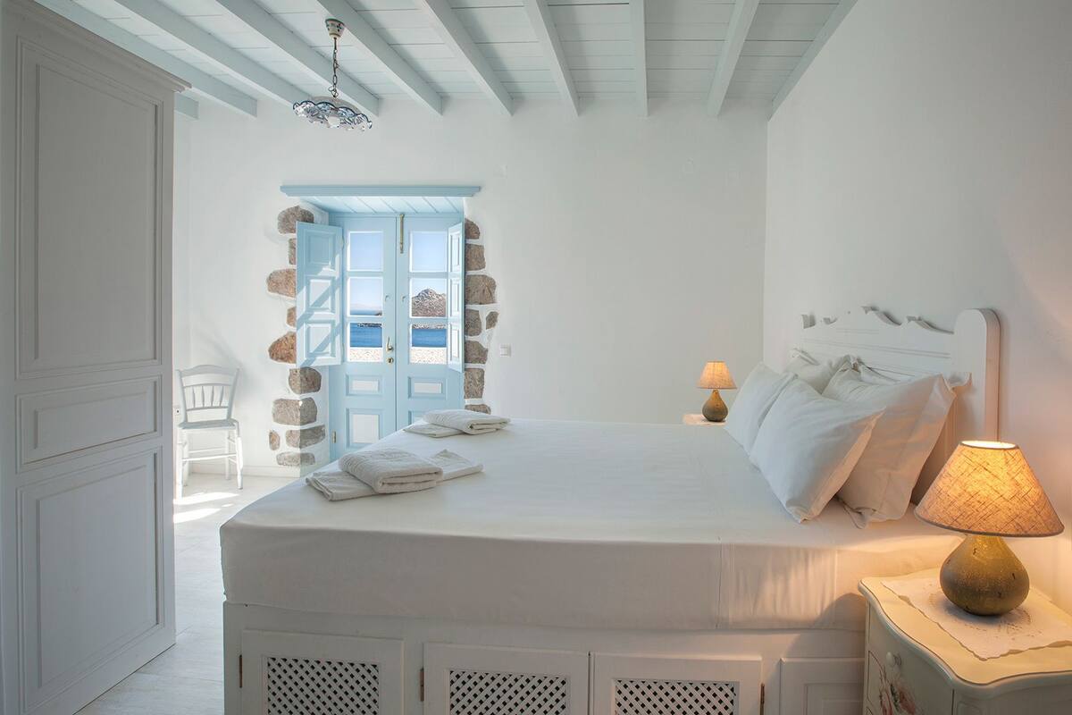 A bedroom with a bed, a nightstand with a lamp, a chair, and a stone wall with a door leading to a balcony.