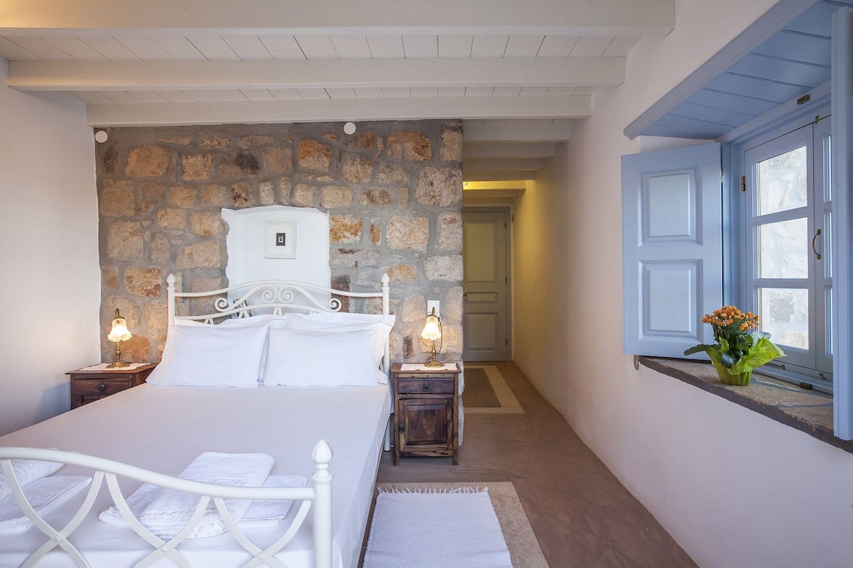 A bedroom with a stone wall, a bed with white linens, two bedside tables with lamps, and a window with blue shutters.