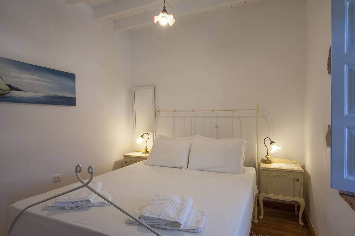 A neatly made bed with white linens, two bedside tables with lamps, a painting on the wall, and a door with a simple design.