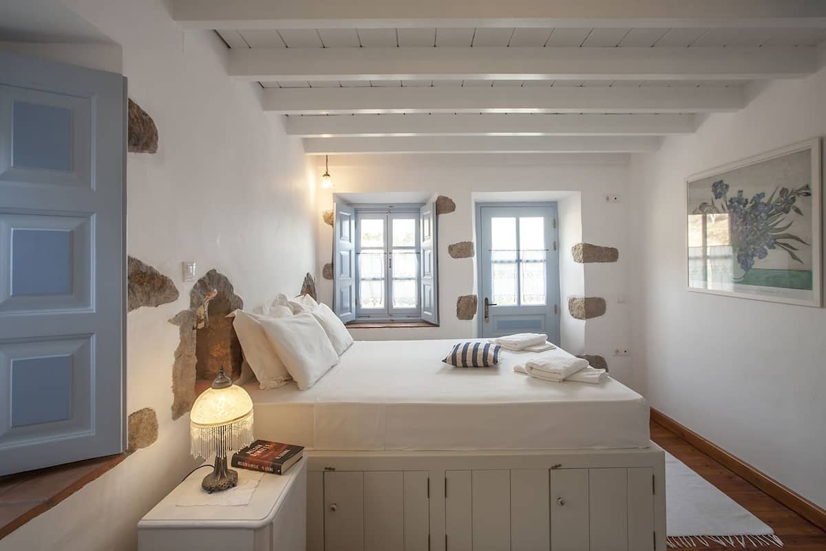 A bedroom with a bed, a nightstand with a lamp, a door, and a window.