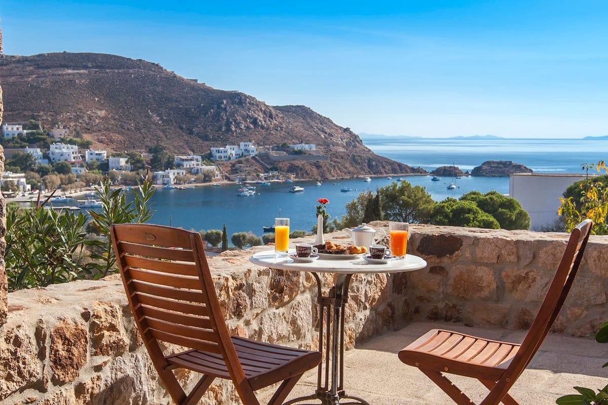 A table with a view of a coastal town, two wooden chairs, and a drink.