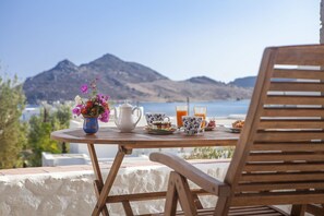A wooden table with a white teapot, a blue and white floral cup, two glasses of orange juice, a plate with pastries, and a vase with flowers. - Theologos Houses (Patmos)