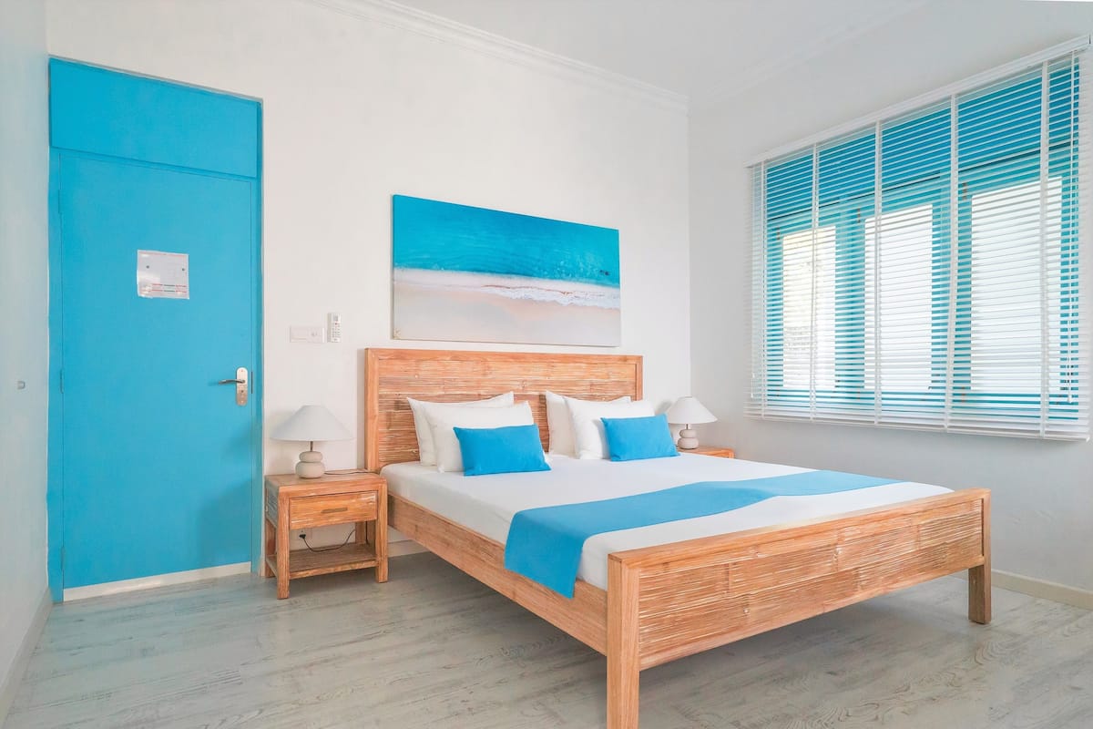 A bedroom with a wooden bed, a blue door, a nightstand with a lamp, and a window with blinds.
