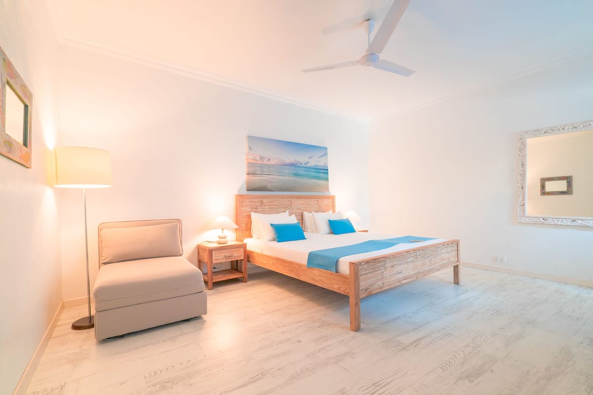 A bedroom with a bed, bedside table, lamp, chair, ceiling fan, and a painting on the wall.