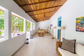 A spacious room with a high ceiling, a dining area with a wooden table and chairs, a sofa with a striped pillow, and a view of greenery outside.