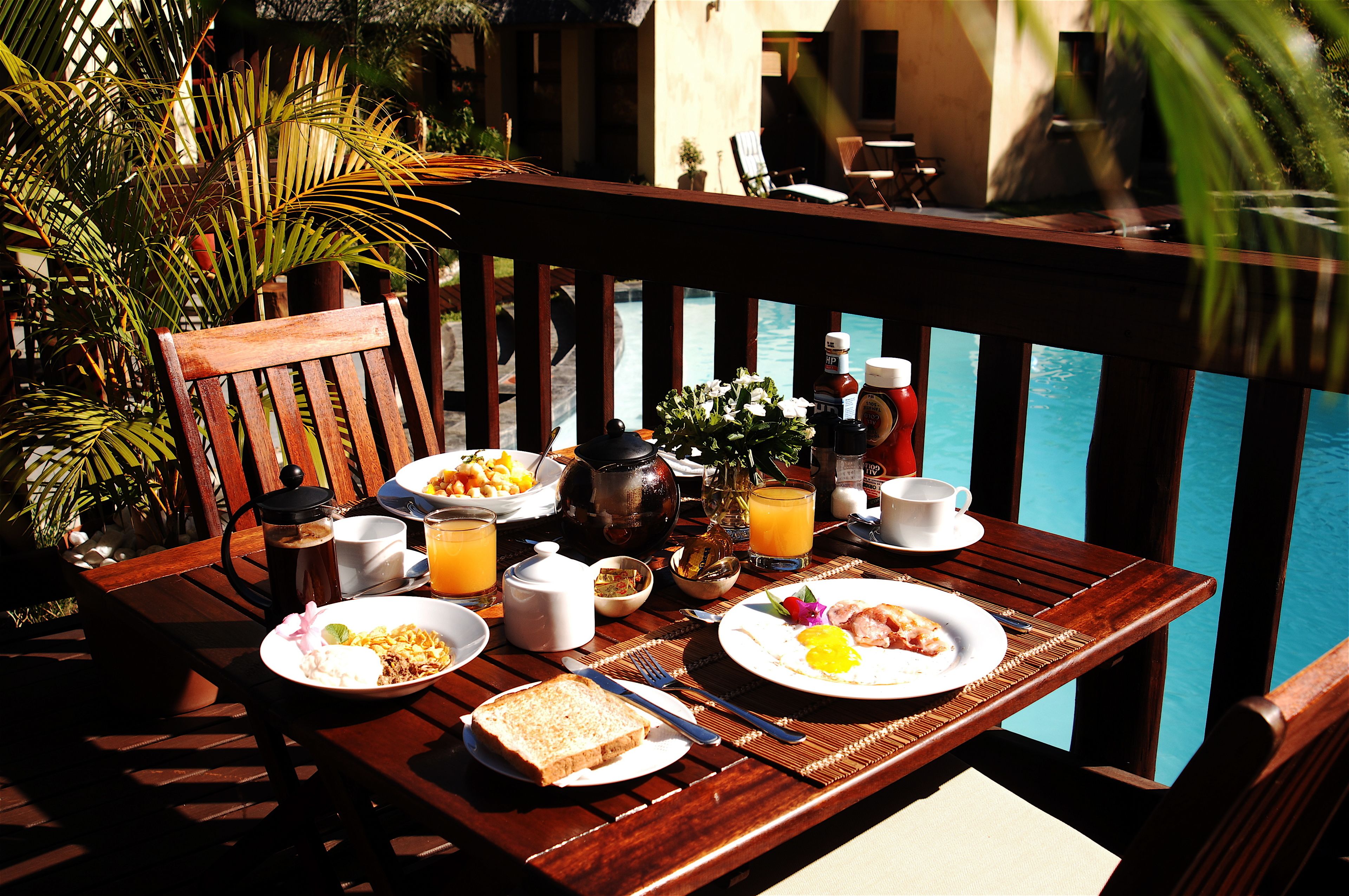 A wooden table with breakfast items, a view of a pool, and a chair.