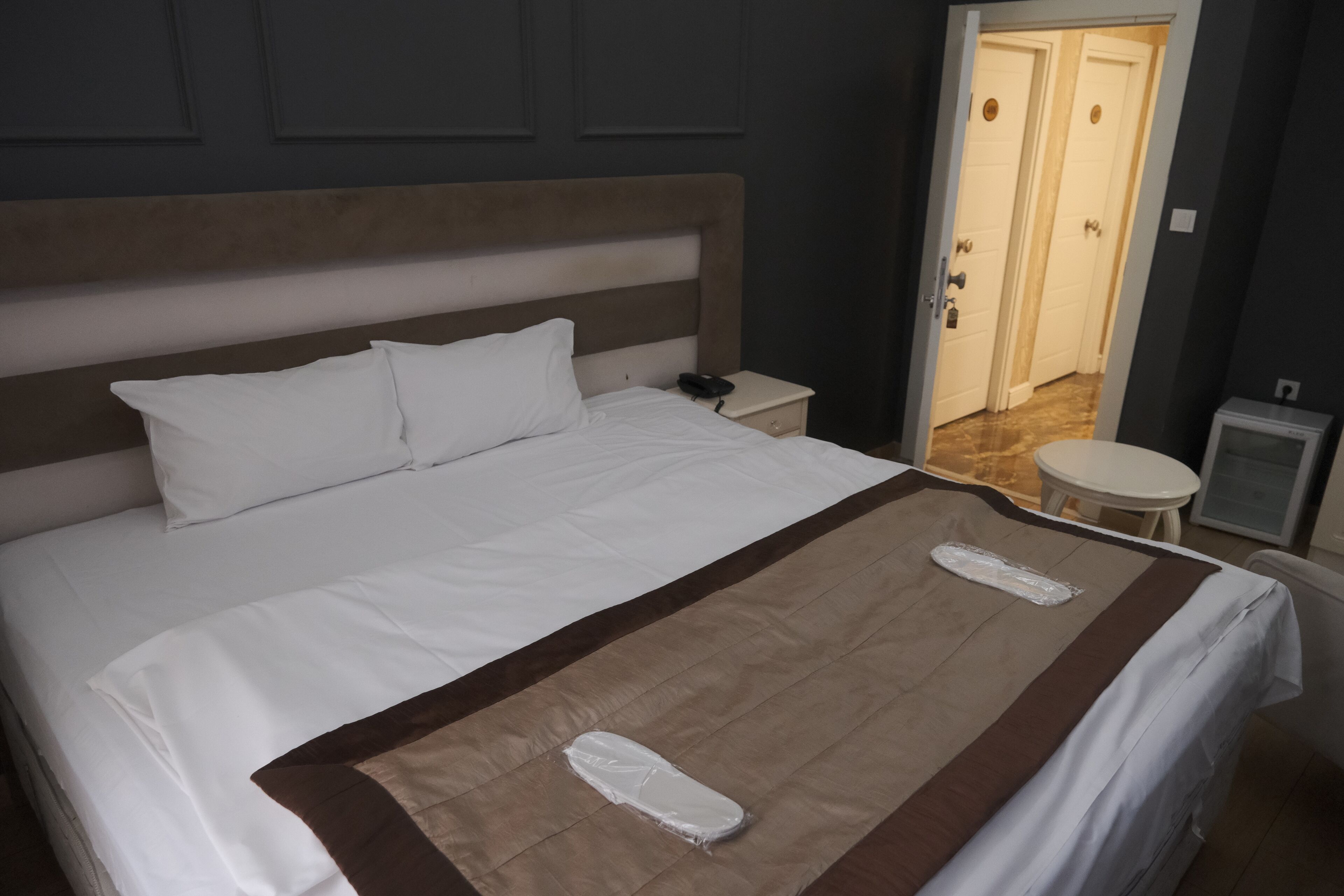 A neatly made bed with white linens and a brown accent, a bedside table with a phone, and a small round table in a hotel room with dark walls and a wooden headboard.