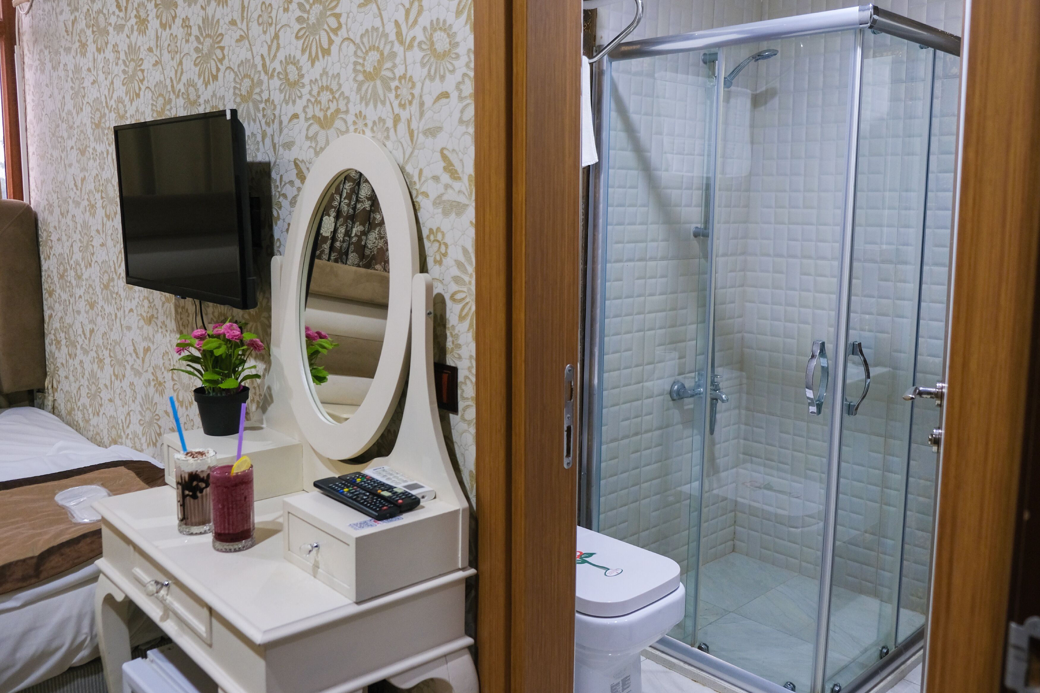 Een hotelbadkamer met een douche, toilet, wastafel met spiegel, televisie en nachtkastje met een plant en een drankje.