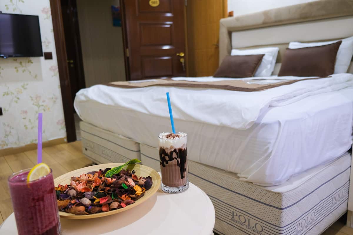 A neatly made bed with white linens and brown pillows, a glass of chocolate milkshake with a blue straw, and a bowl of assorted fruit on a round table.