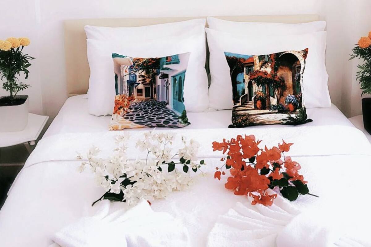Una cama bien hecha con sábanas blancas, dos almohadones decorativos con un diseño de paisaje urbano y un ramo de flores blancas y rojas sobre la cama.