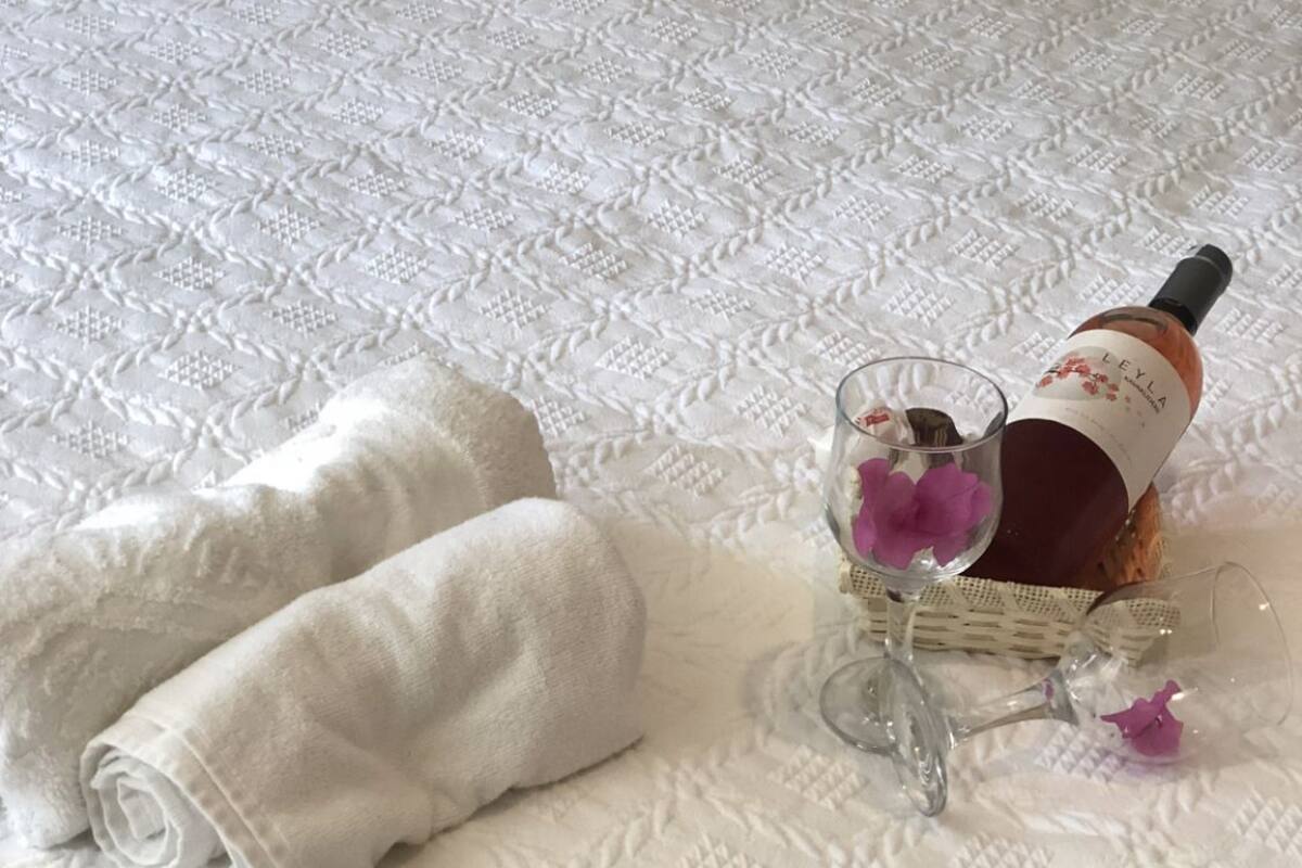Una cama bien hecha con un edredón blanco texturizado, una toalla blanca enrollada, una botella de vino y una copa con una flor morada.