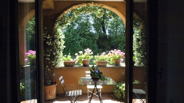 Eine Terrasse im Freien mit einem Glastisch und zwei Stühlen, umgeben von Kübelpflanzen und einer Steinmauer.