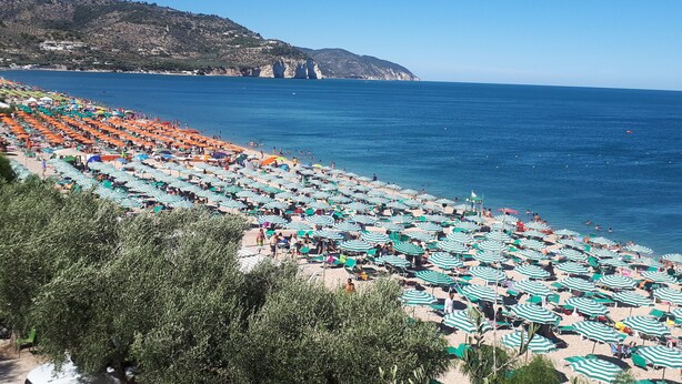 Una playa con hileras de sombrillas y sillas de descanso, un cielo azul claro y montañas al fondo.