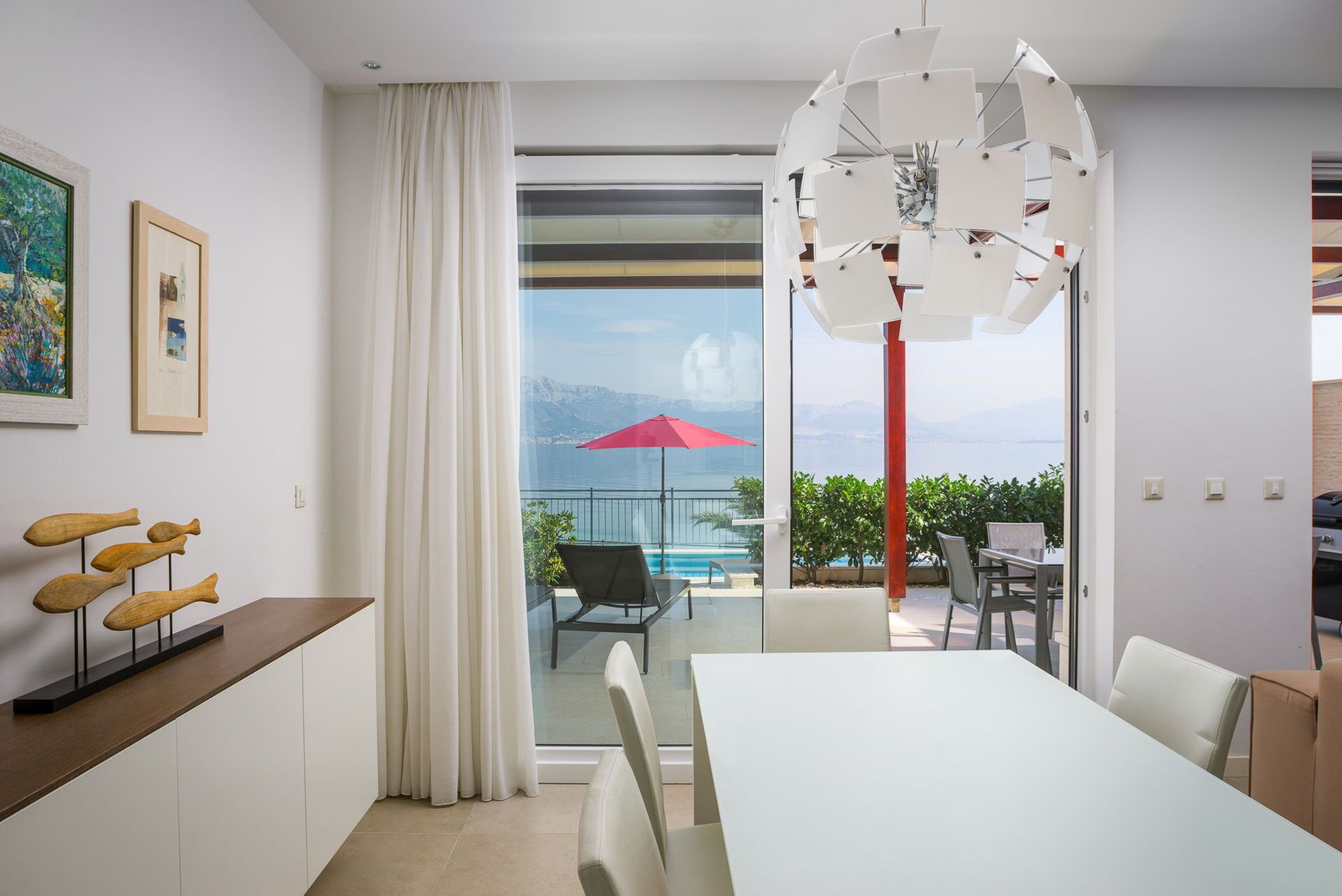 A modern living room with a large window, a dining area, and a view of a pool and mountains.
