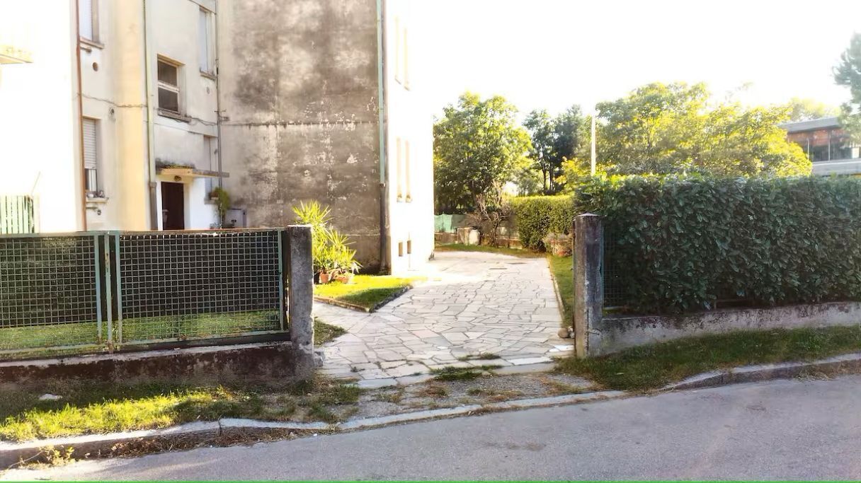 Un chemin pavé mène à un bâtiment doté d’une grille en métal et bordé d’une haie.