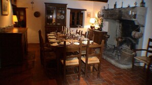 Un comedor con una mesa de madera dispuesta para seis personas, rodeada de sillas. Hay una chimenea con un hogar de piedra y un reloj en la pared.