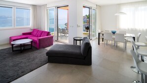 Ein modernes Wohnzimmer mit einem rosa Sofa, einem grauen Sofa, einem Esstisch mit Stühlen und einem Balkon mit Meerblick.