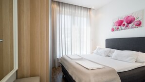Ein Hotelzimmer mit Bett, Nachttisch, Fenster mit durchsichtigen Vorhängen und einem floralen Bild an der Wand.