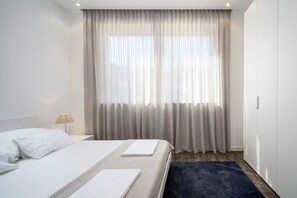 A hotel room with a bed, a nightstand, a lamp, and a window with sheer curtains.