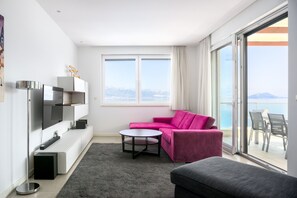 A modern living room with a pink sofa, a grey ottoman, a flat-screen TV, and a large window offering a view of the sea.