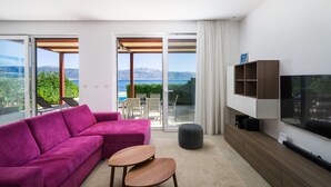 A modern living room with a pink sofa, a wooden coffee table, and a TV on a wooden cabinet. There are sliding glass doors leading to a balcony with a view of mountains and the sea. - Beachfront Villa Lemonade with Pool III (Trogir)