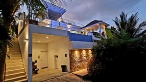 A modern two-story house with a staircase leading to the upper level, a covered outdoor area, and a stone wall. - Villa Paradise with pool and sea view (Ko Samui)