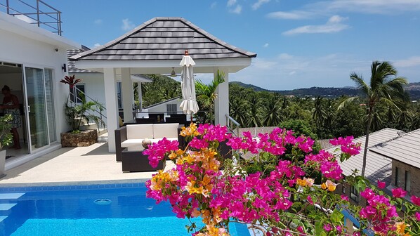 Un espace piscine extérieur moderne doté d’un coin salon abrité, offrant une vue dégagée sur le paysage environnant et agrémenté de magnifiques fleurs de bougainvillier aux couleurs vives.