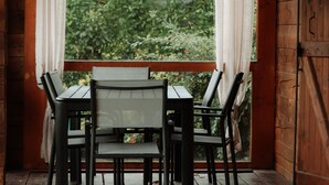 Een houten terras met een tafel en stoelen, uitzicht op groen door een raam, en een houten deur.