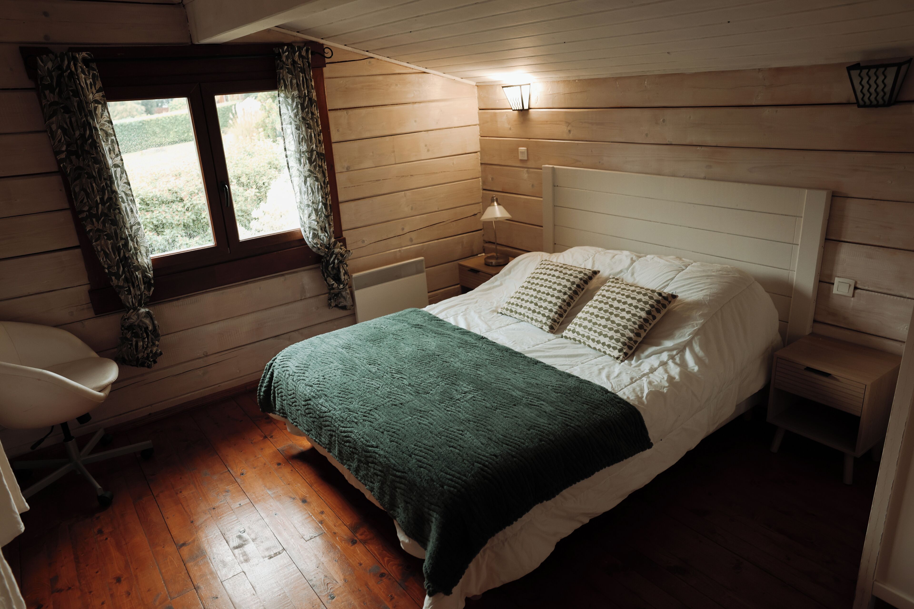 Une pièce chaleureuse avec un plancher en bois, un lit recouvert d’une couverture verte, une lampe de chevet et une fenêtre avec des rideaux.
