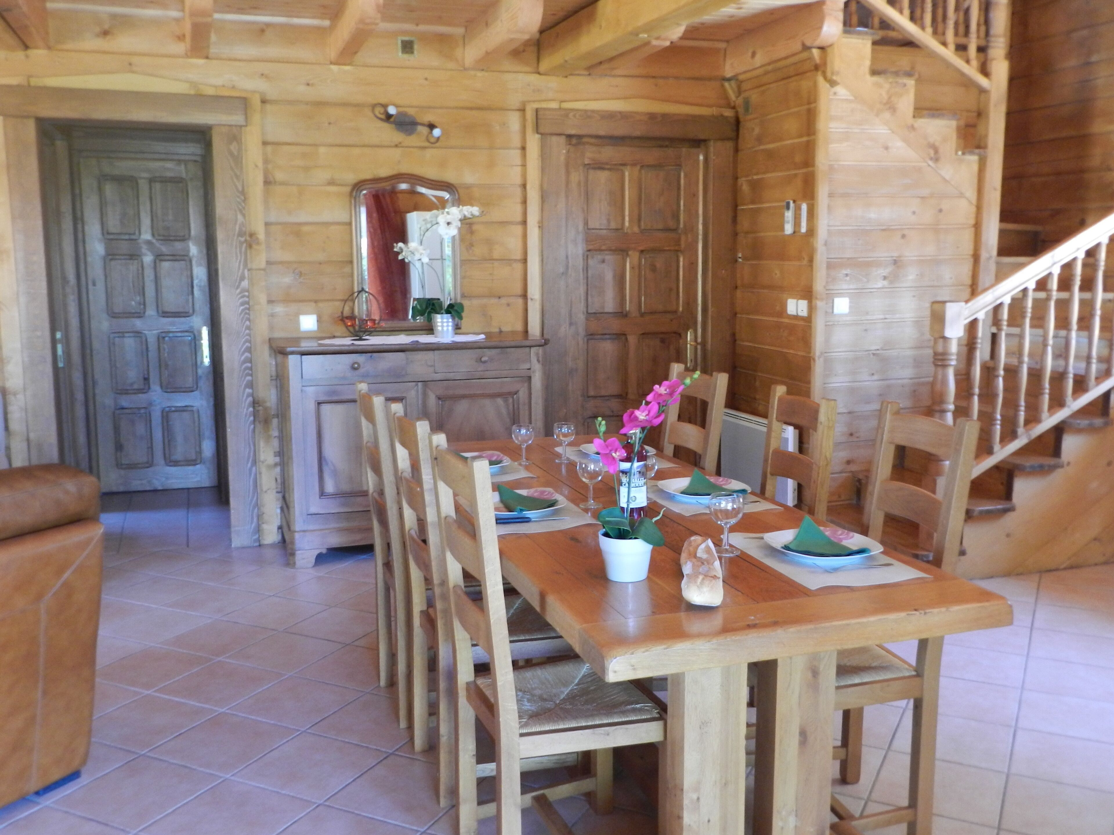 Een houten eetkamer met een lange tafel gedekt voor zes personen, omringd door houten stoelen. Een houten trap is rechts zichtbaar.