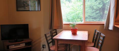 A room with a television on a wooden stand, a small table with a potted plant, and a window with curtains.