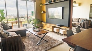 A modern living room with a large TV, comfortable seating, a dining area, and a view of the city. - Padgett Place - Deluxe Suites (Cebu City)