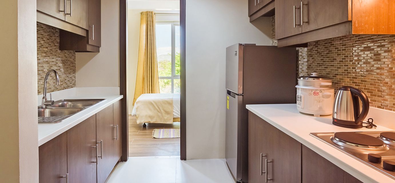 A compact kitchen with a refrigerator, coffee maker, and kettle. A bed is visible through an open door in the background.