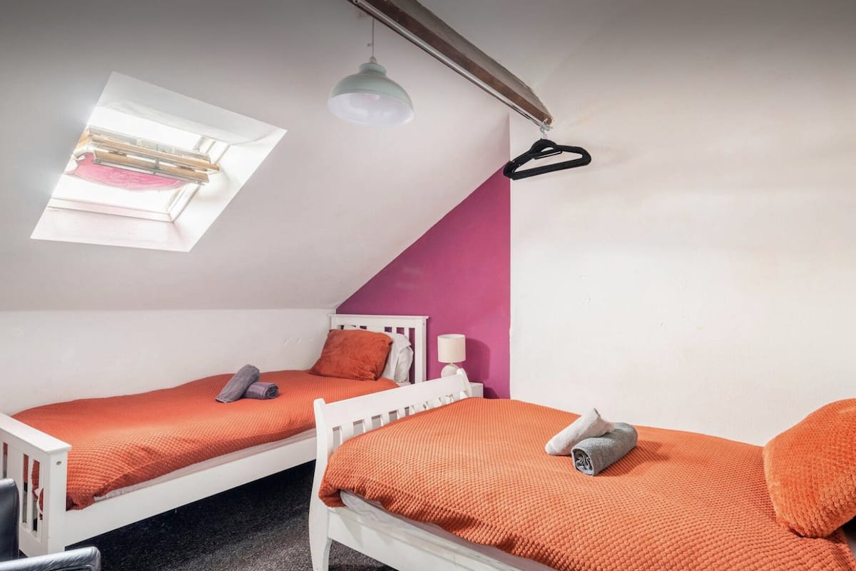 Deux lits simples avec une literie orange, une tête de lit blanche, une table de chevet blanche avec une lampe, et une fenêtre avec un store blanc.