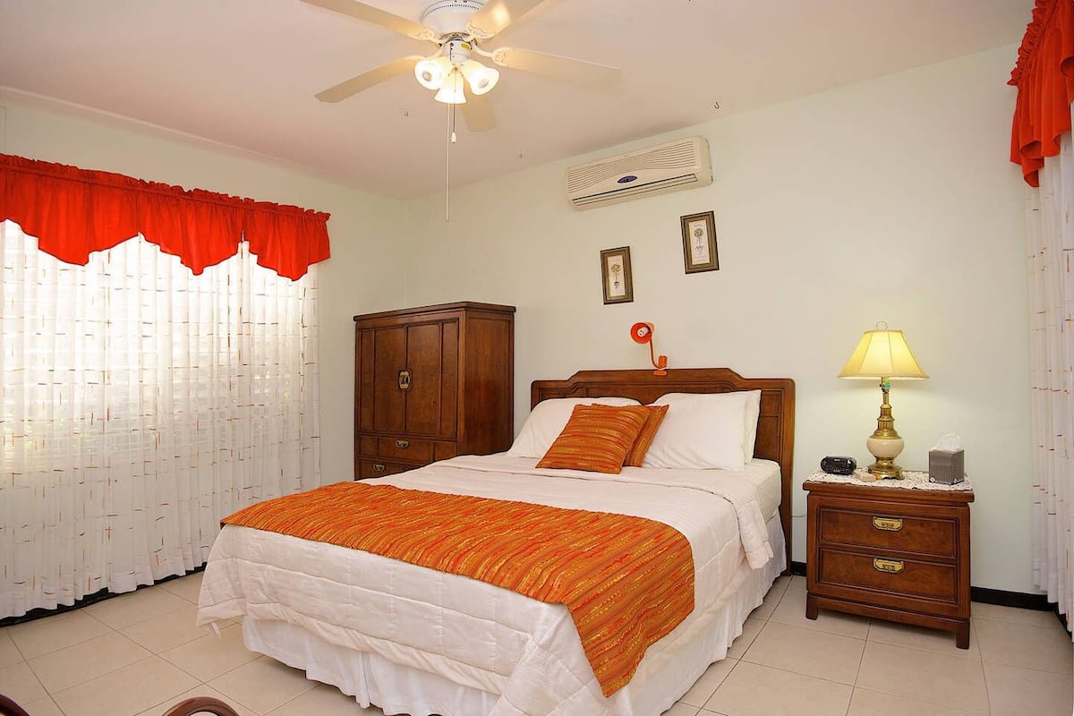 A bedroom with a bed, a nightstand, a wardrobe, a ceiling fan, and an air conditioning unit.