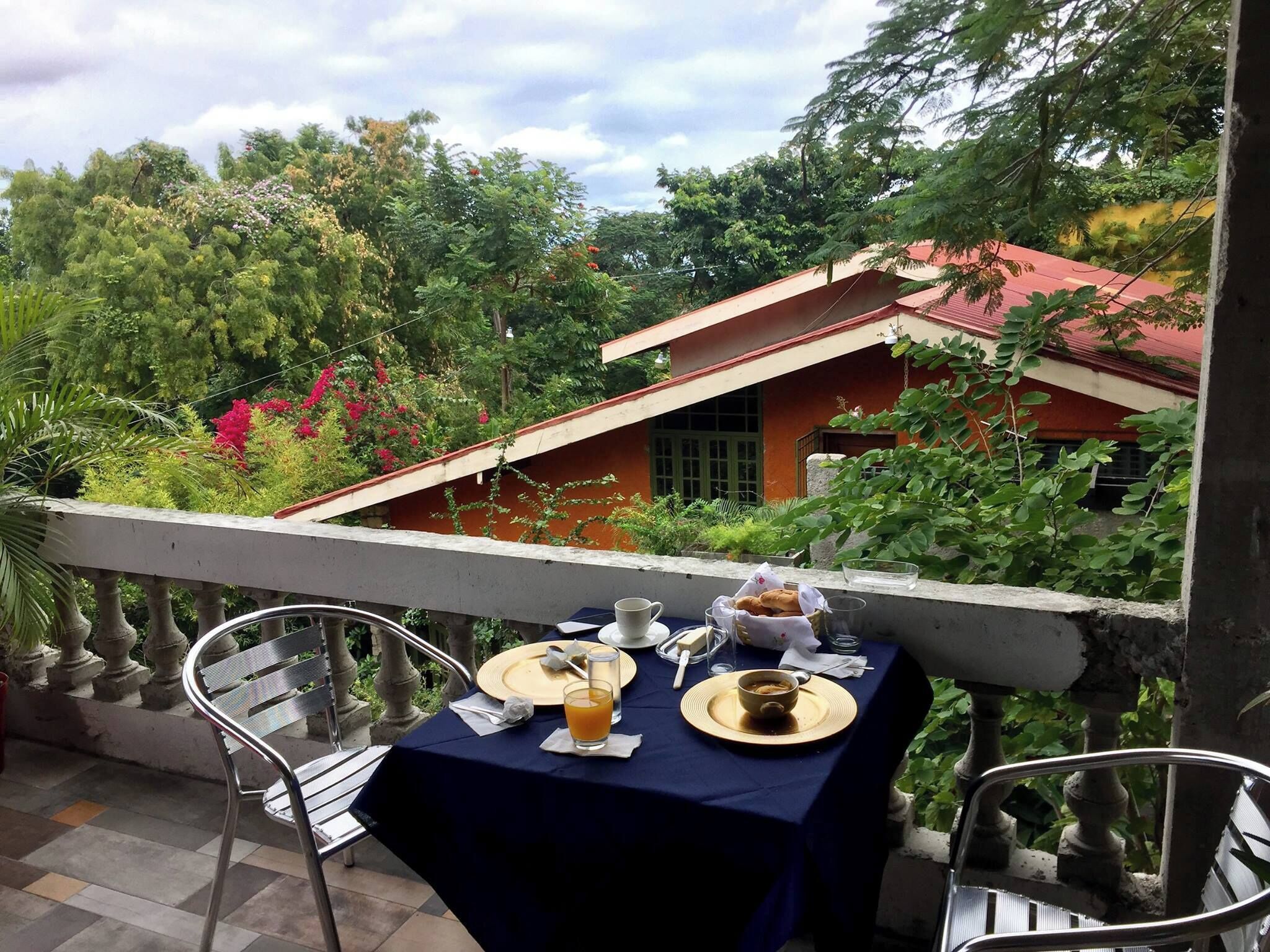 Une table dressée pour le petit-déjeuner sur un balcon, avec vue sur une maison et une végétation luxuriante.