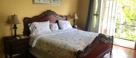 A bedroom with a wooden bed, floral bedding, two bedside lamps, a nightstand, and a window with a view of greenery.