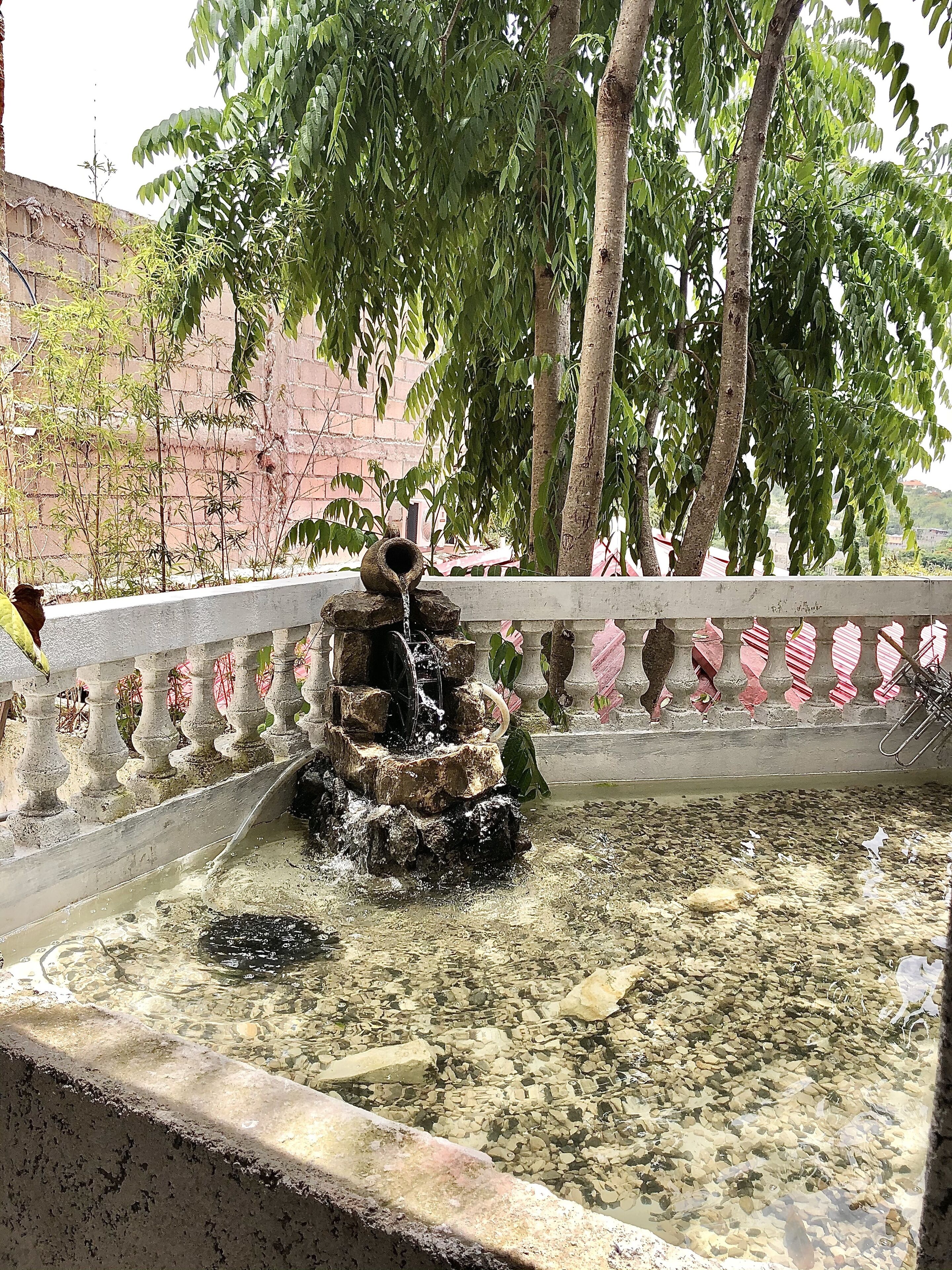 A water feature with a cascading stone structure in a courtyard, surrounded by trees and a brick wall.