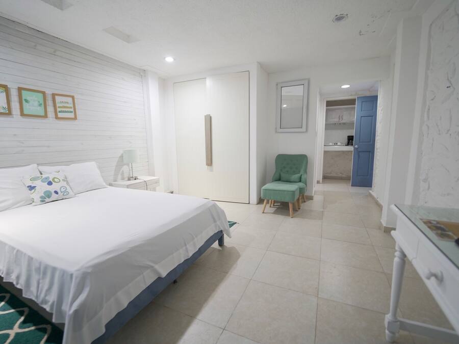 Quarto com cama, mesa de cabeceira, cadeira e escrivaninha. Há uma porta que dá para um banheiro.
