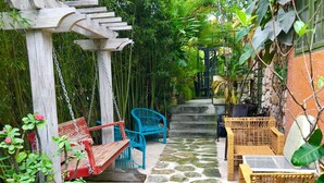 Un chemin de pierres mène à une pergola en bois agrémentée d’une balançoire, le tout étant entouré d’une végétation luxuriante et de meubles en osier.