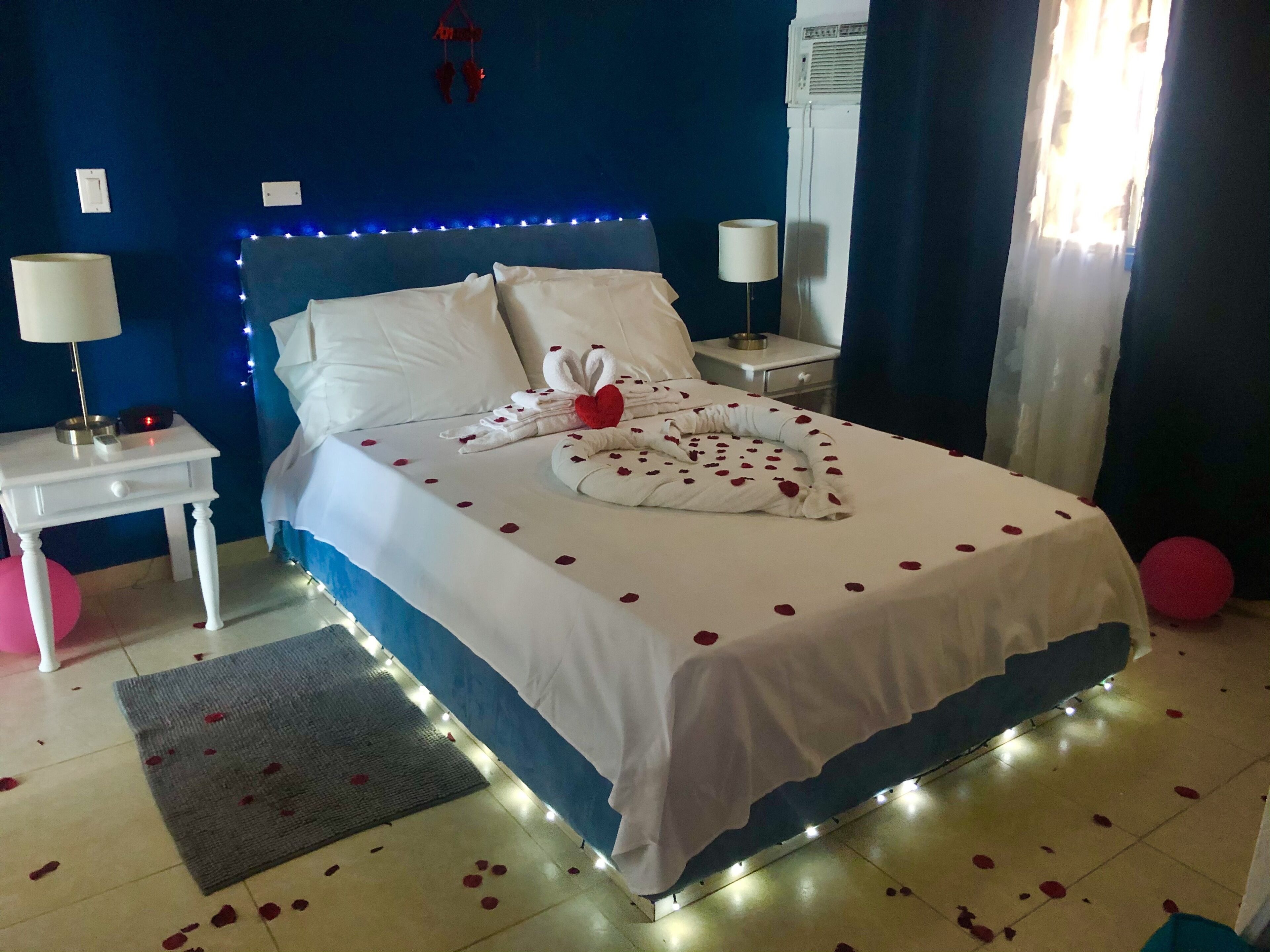 Een net opgemaakt bed met witte lakens en rozenblaadjes, een bedlampje en een klein tafeltje met een lamp en een rood object.