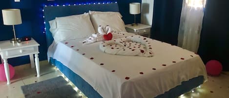 A neatly made bed with white linens and rose petals, a bedside lamp, and a small table with a lamp and a red object.
