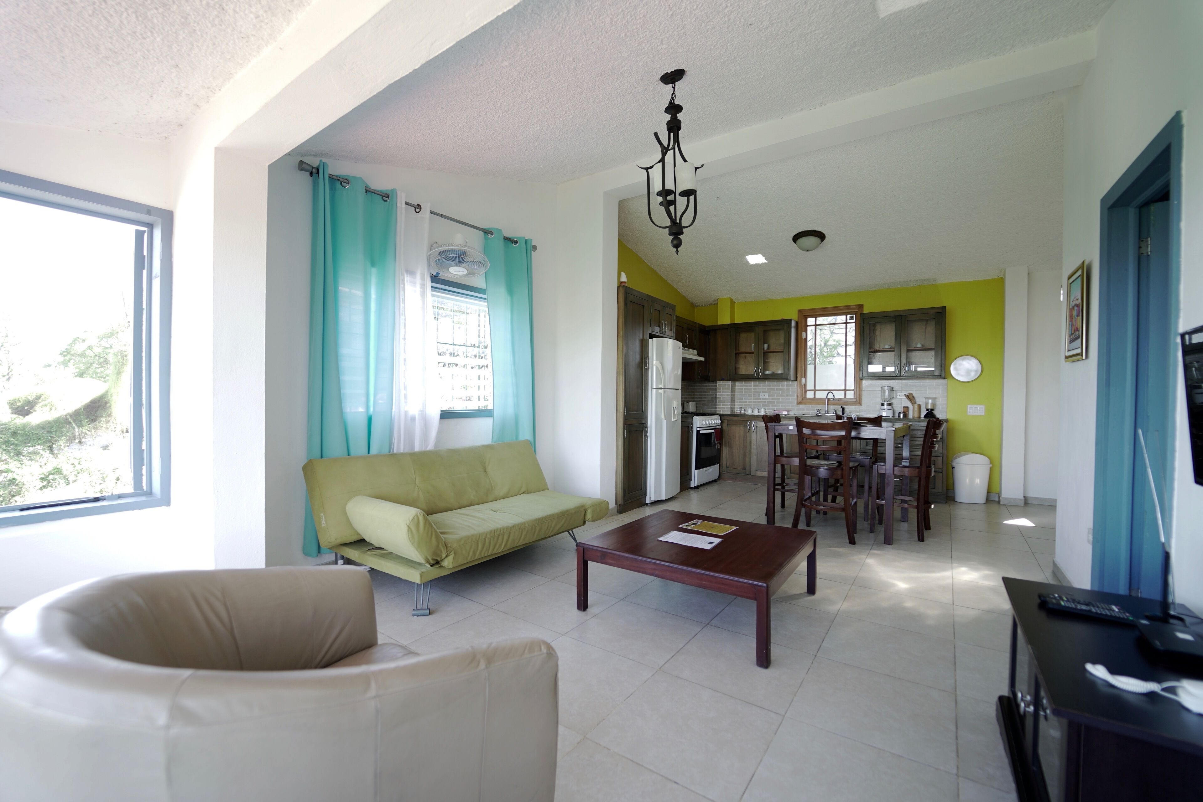 A living room with a sofa, a coffee table, and a dining area with chairs and a table. There is a kitchen area with cabinets and a refrigerator.