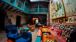 A cozy interior with a high ceiling, wooden beams, and a stone wall. There are blue chairs, wooden tables, and a chessboard. - Nao Victoria Hostel (Cusco)