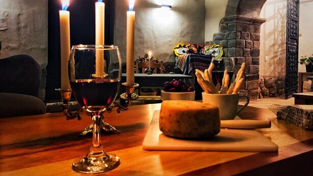 A candlelit room with a wooden table featuring a glass of red wine, a cheese platter, and a candle holder. - Nao Victoria Hostel (Cusco)