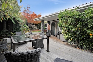 A patio with wicker furniture, a glass-top table, and a view of a house with a covered outdoor area and a tree with oranges. - Leinster Villa (Christchurch)