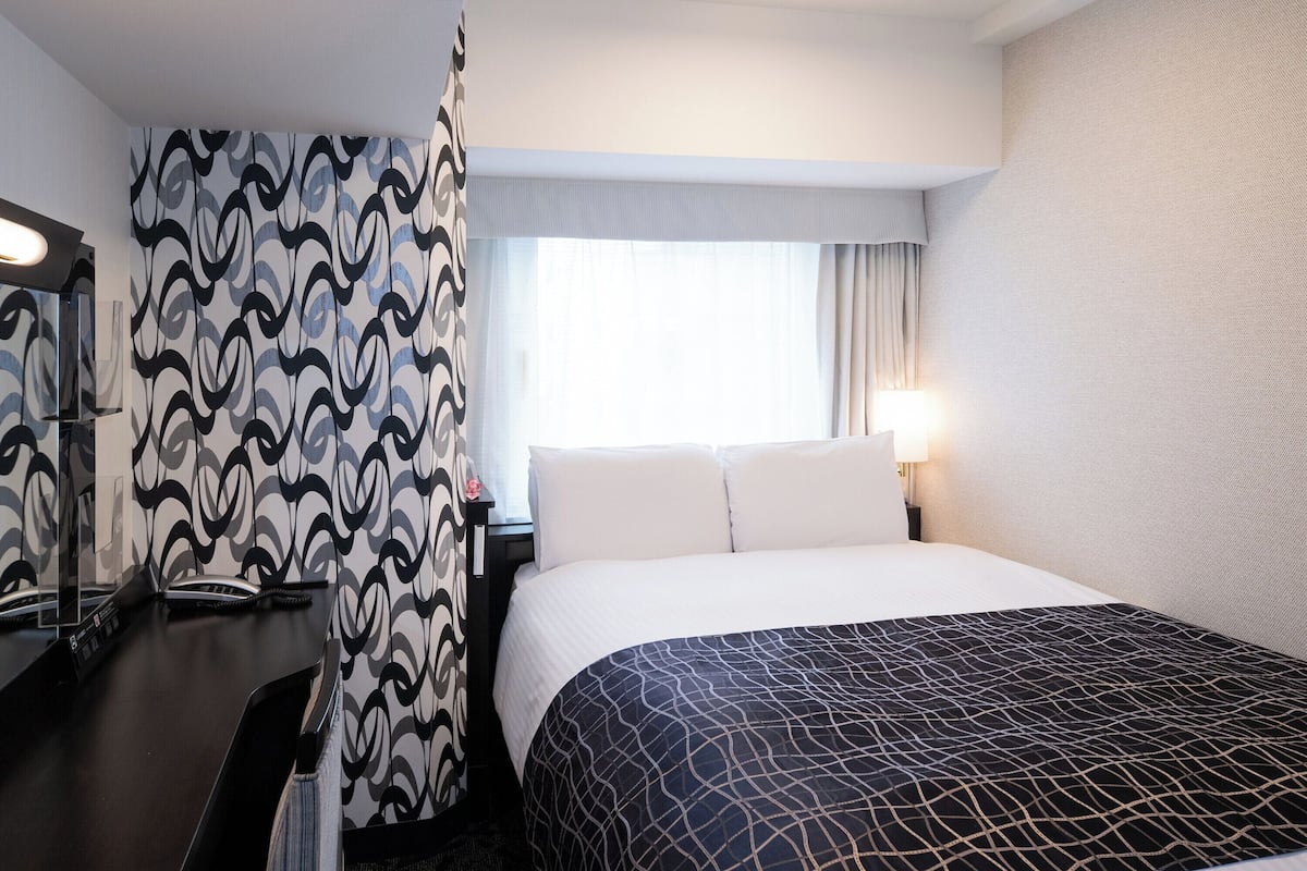 A hotel room with a bed, a desk, a telephone, and a window with curtains.