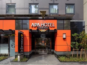 The exterior of APA Hotel with a modern design, featuring a prominent orange facade, large windows, and a clear entrance.
